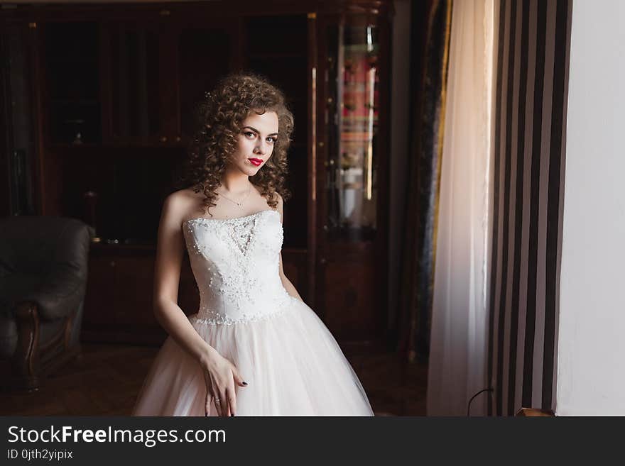 An Elegant Girl Standing By The Window Dressed In A Wedding Dress She Has Curly Hair