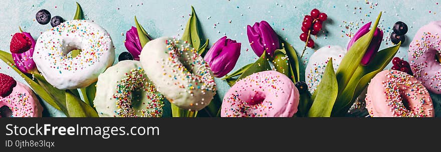 Sweet and colourful doughnuts with sprinkles, purple tulips and berries falling or flying in motion against blue pastel background. Border with space for text