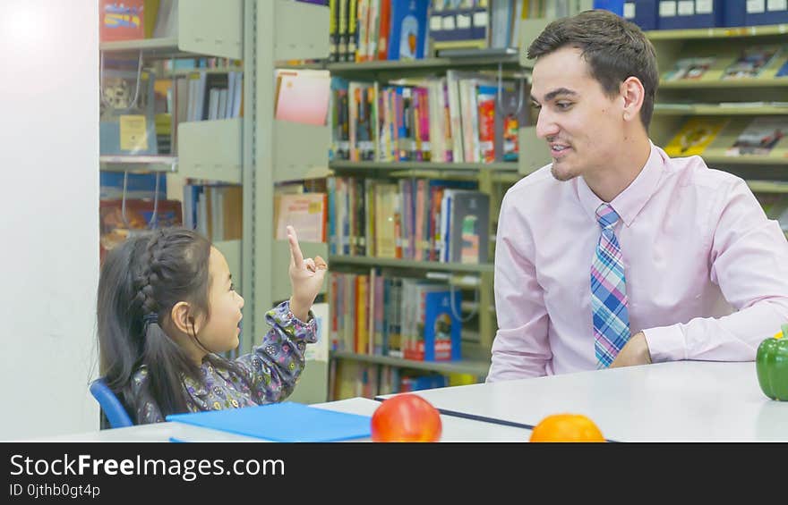 Smiley caucasian teacher and grouping of asian kids student learning and talking at white table and color book with bookshelf background