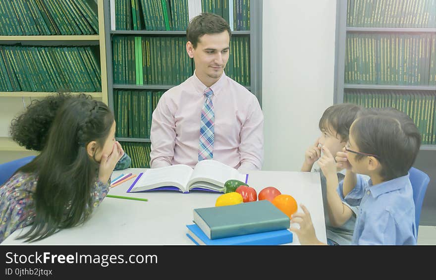 Smiley caucasian teacher and grouping of asian kids student learning and talking at white table and color book with bookshelf background