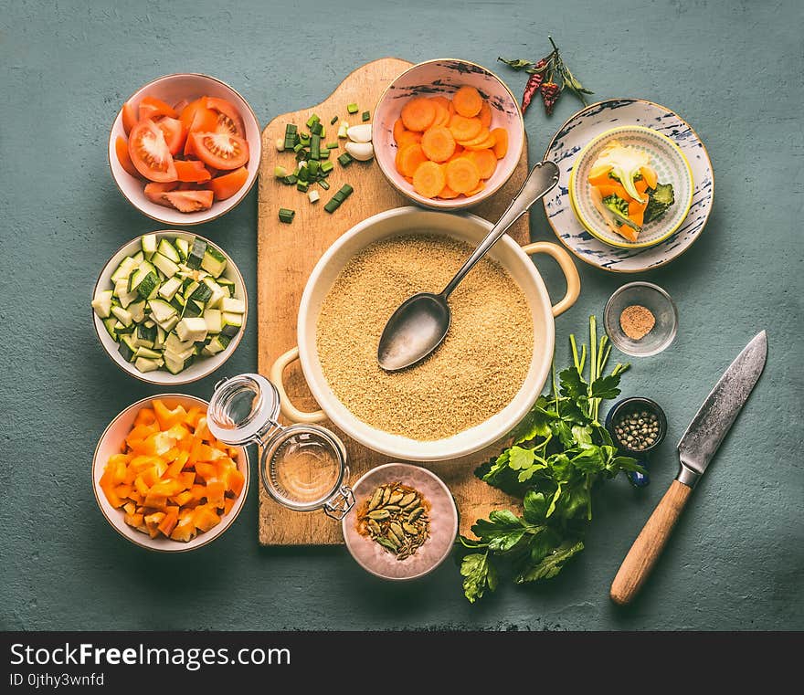 Couscous and vegetarian cooking ingredients in bowls on cutting board with spoon , gray background, top view