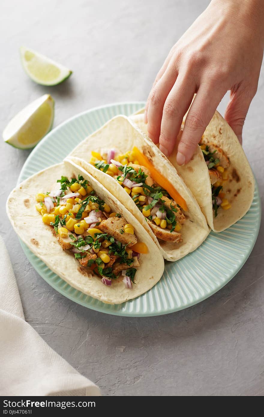 Hand takes taco from the blue plate on the dinner table. Mexican snacks with flatbread, corn, chicken meat and parsley.