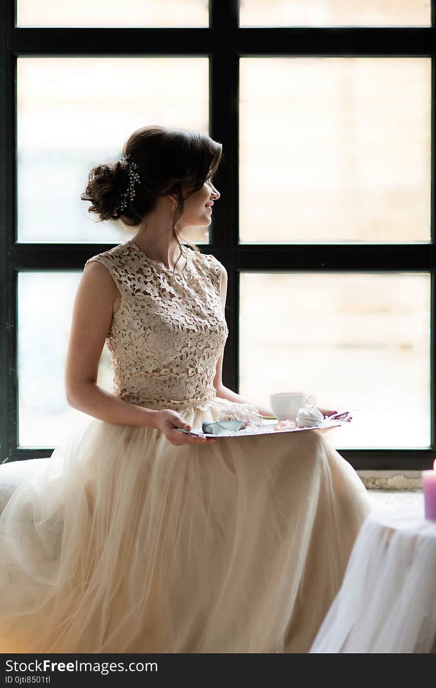 Young gorgeous woman wearing sleeveless evening dress. She is waiting for her boyfriend sitting in front of window with tray of sweets and coffee.