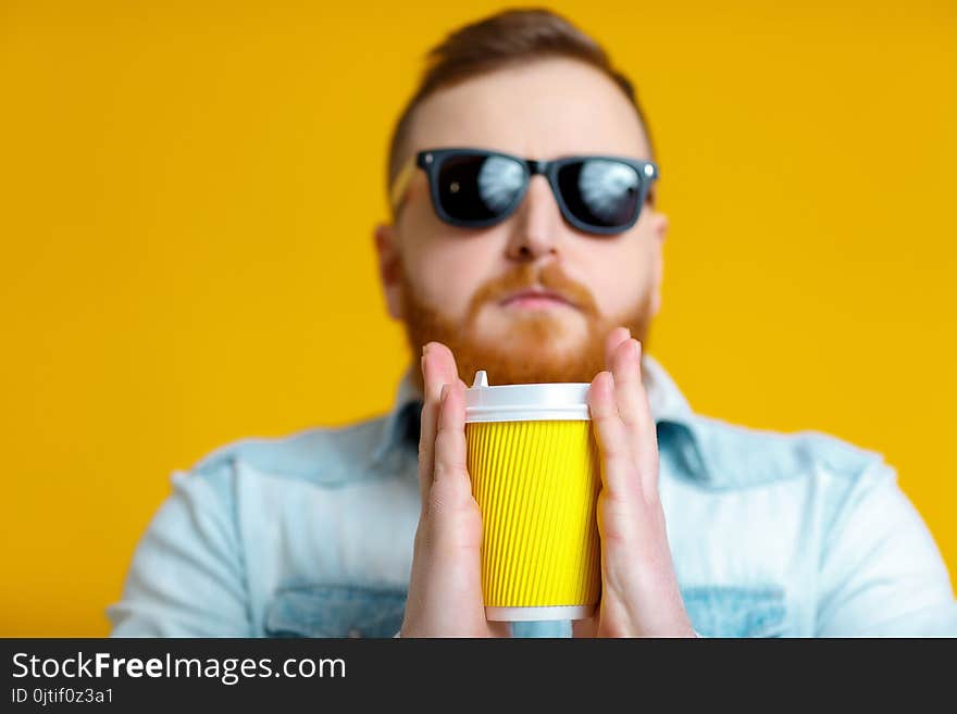 Red beard man in sunglasses and denim shirt holding paper cup with coffee on yellow background. Red beard man in sunglasses and denim shirt holding paper cup with coffee on yellow background.