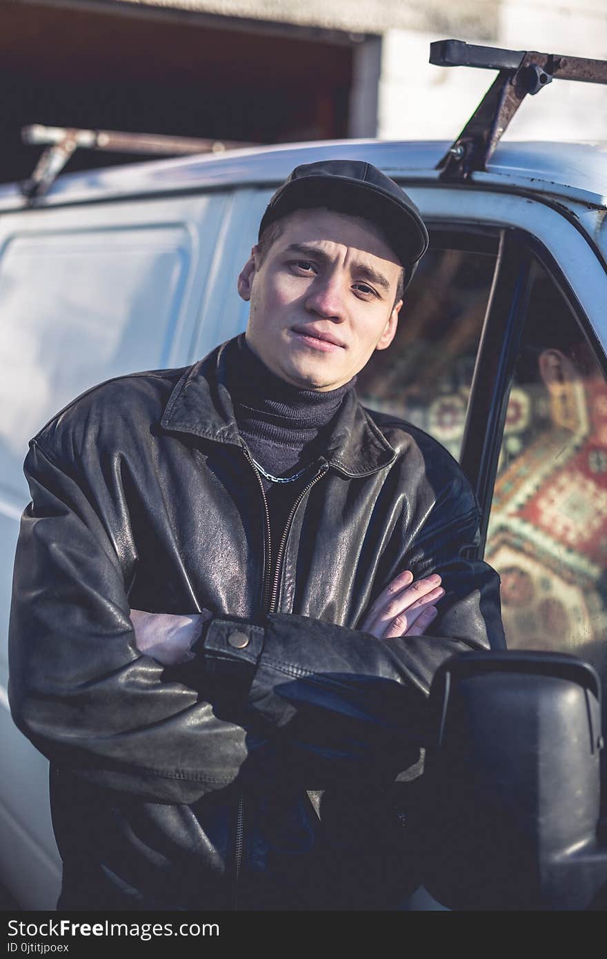 A young guy of criminal appearance in a black leather jacket stands near an old white van.