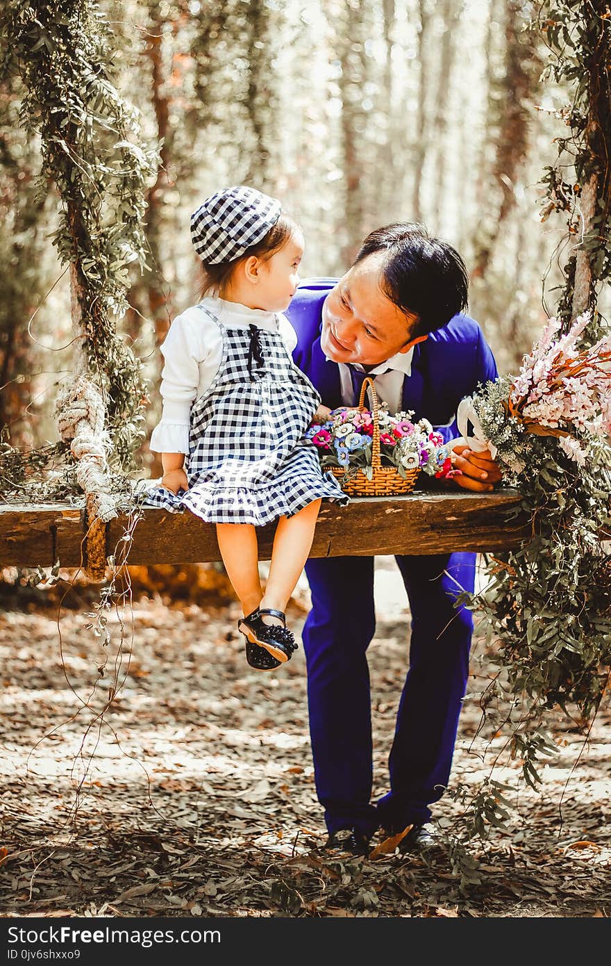 Selective Focus and Color Photography of Man Looking at Her Girl Sitting on Garden Swing White Holding Bouquet of Flower in Brown Wicker Basket