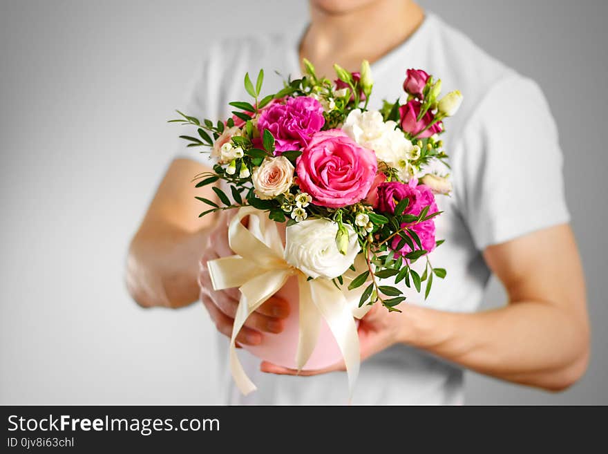 Man in white t shirt holding in hand rich gift bouquet. Composition of flowers in a pink hatbox. Tied with wide white ribbon and bow.