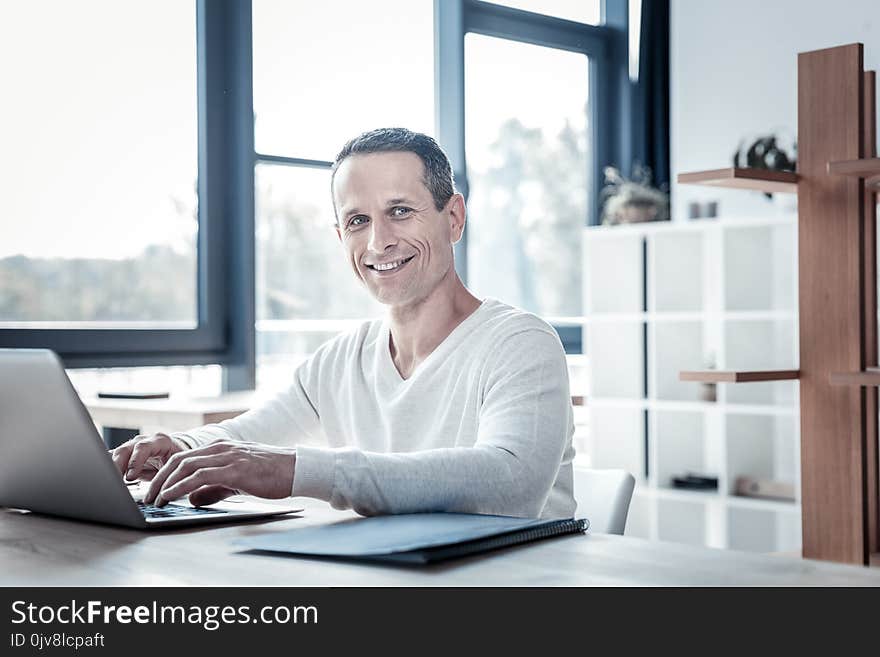 Adore my work. Successful qualified satisfied man sitting by the table smiling and using the laptop. Adore my work. Successful qualified satisfied man sitting by the table smiling and using the laptop.