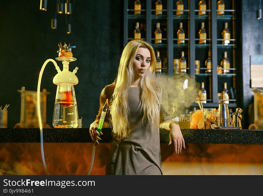 Young Woman Smoking A Hookah Behind The Bar. Cloud Of Smoke. Beauty Portrait Of Young Woman Enjoying The Hookah