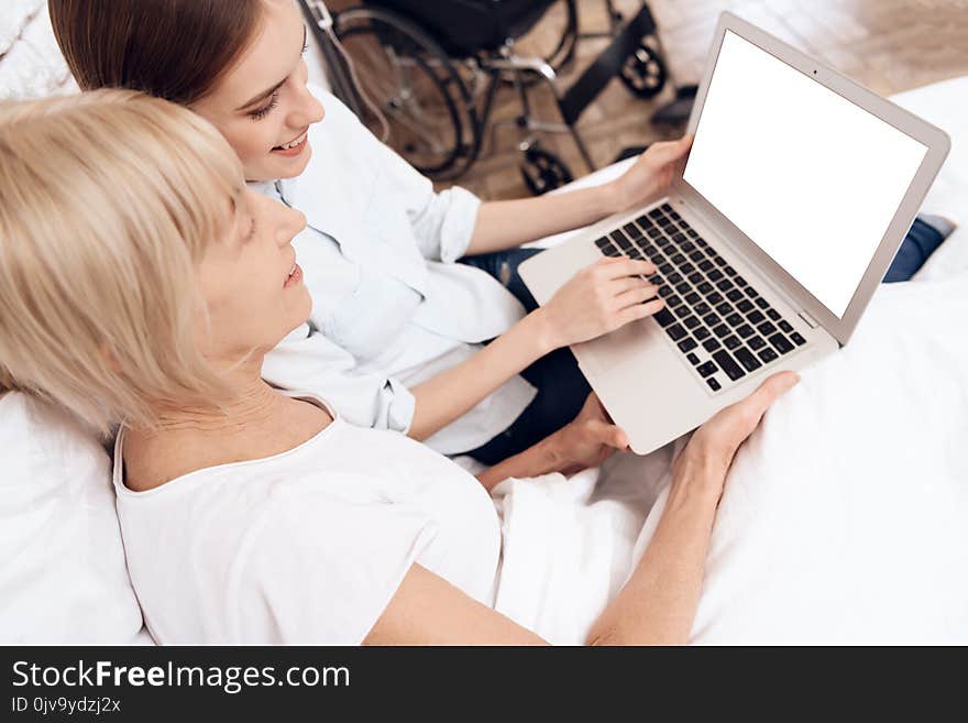 Girl is caring for elderly women in bed at home. They are using laptop. They are happy.