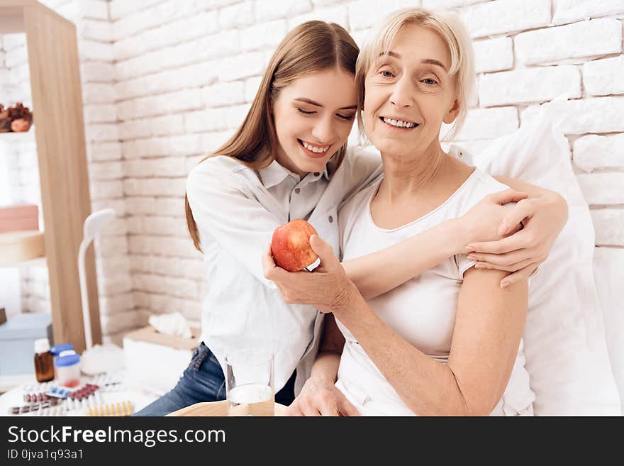 Girl is nursing elderly women in bed at home. They are embracing. Woman is holding apple.