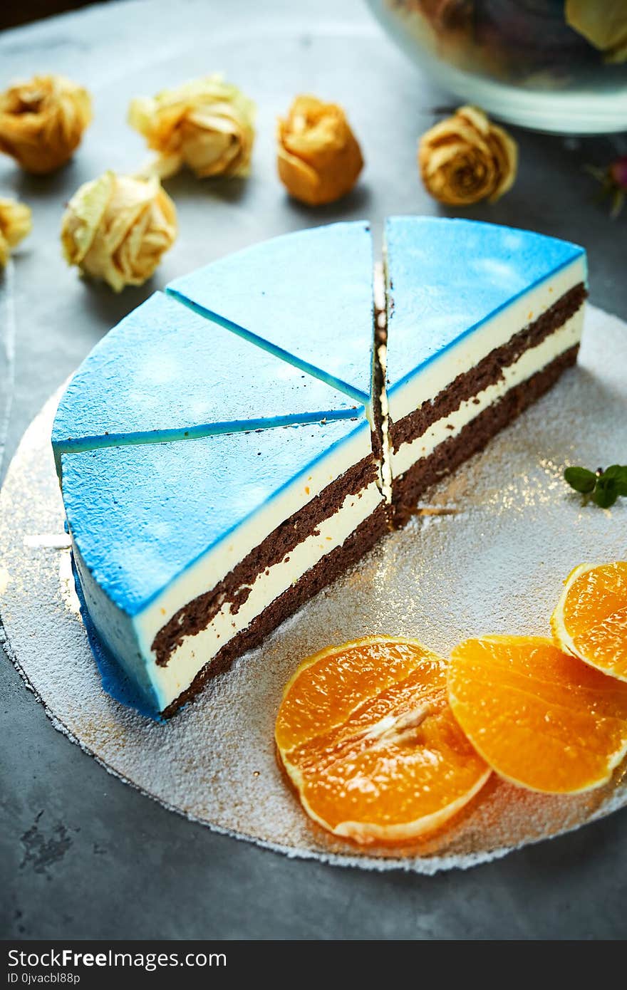 Top view of composition of half of cutted mousse cake with blue glaze, sliced oranges, sugar powder and dried roses on a dark gray concrete background. Top view of composition of half of cutted mousse cake with blue glaze, sliced oranges, sugar powder and dried roses on a dark gray concrete background