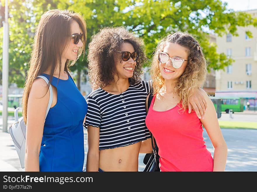 Outdoors portrait of three happy female multiethnic friends in sunglasses. Girls in colourful clothes having a walk in city in summer and having fun, copy space. Urban lifestyle, friendship concept. Outdoors portrait of three happy female multiethnic friends in sunglasses. Girls in colourful clothes having a walk in city in summer and having fun, copy space. Urban lifestyle, friendship concept