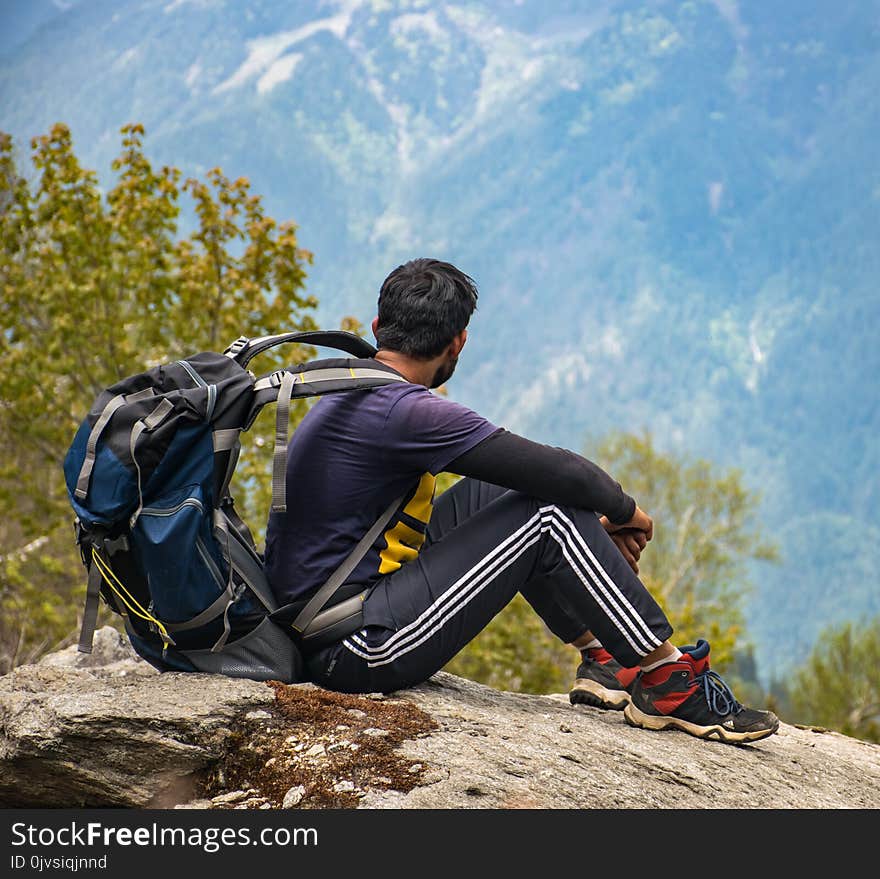 Man in Purple T-shirt With Blue Backpack Sitting on Gray Boulder