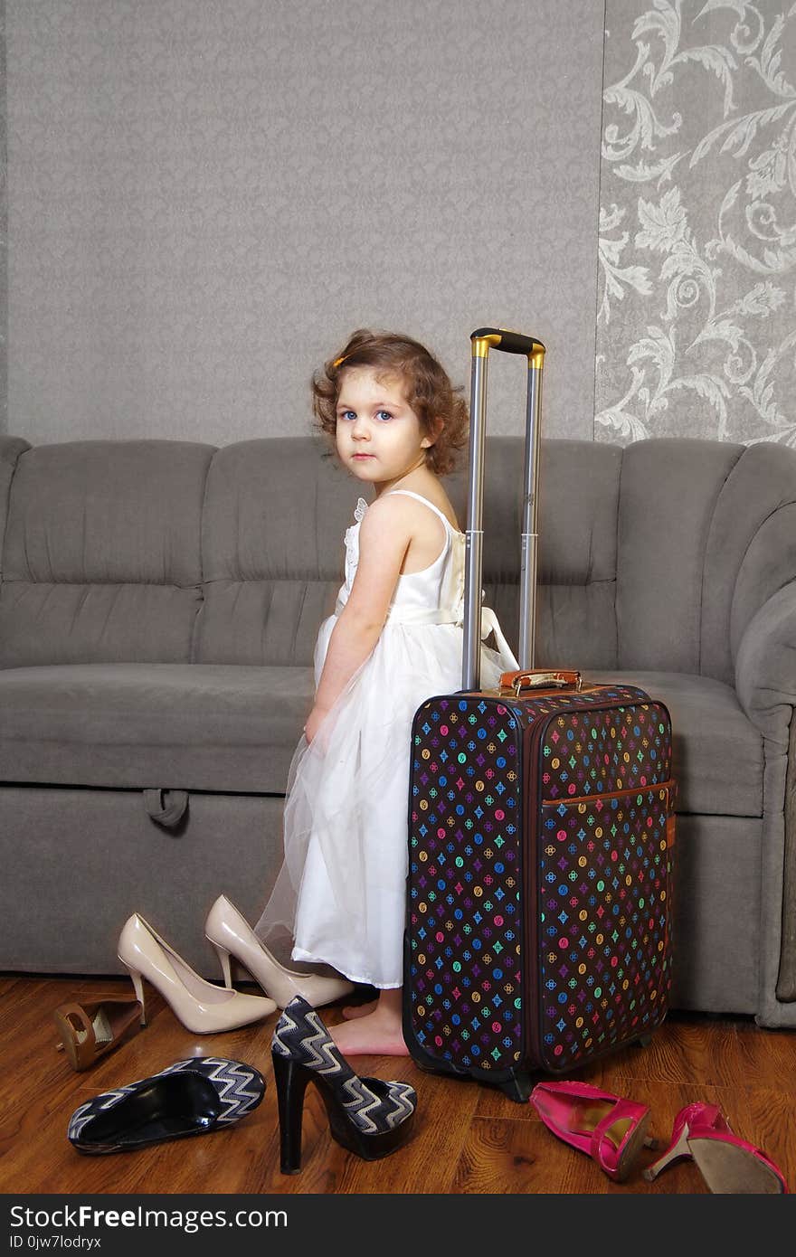 The little girl in a white dress with a suitcase beside her and the adult women`s shoes. The little girl in a white dress with a suitcase beside her and the adult women`s shoes.