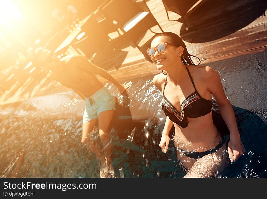 Young happy couple is resting under sun in swimming pool at summer time. Company of friends carefree spend time swimming in pool.