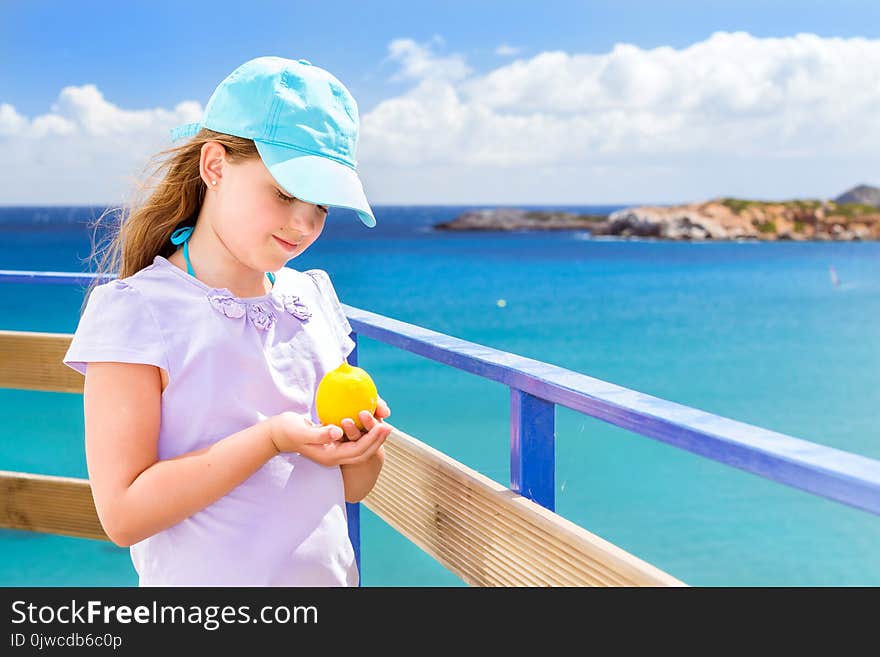 Cute happy girl in a summer dress with a ripe yellow lemon in hand posing against background of ultramarine turquoise Cretan sea. Cliff above Livadi beach in resort village Bali, Crete, Greece. Cute happy girl in a summer dress with a ripe yellow lemon in hand posing against background of ultramarine turquoise Cretan sea. Cliff above Livadi beach in resort village Bali, Crete, Greece