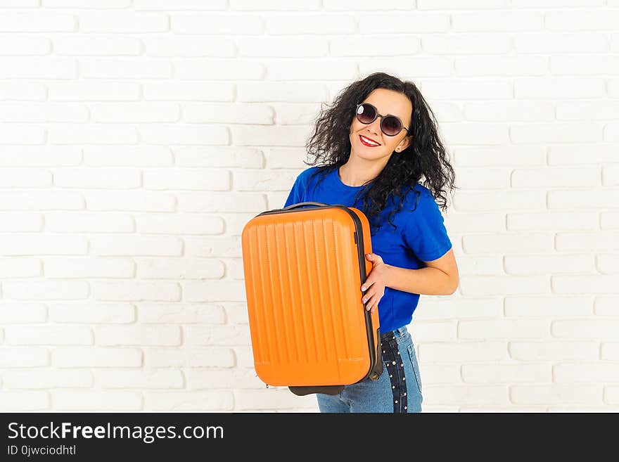 Happy young woman in sunglasses holds modern suitcase on white brick wall.