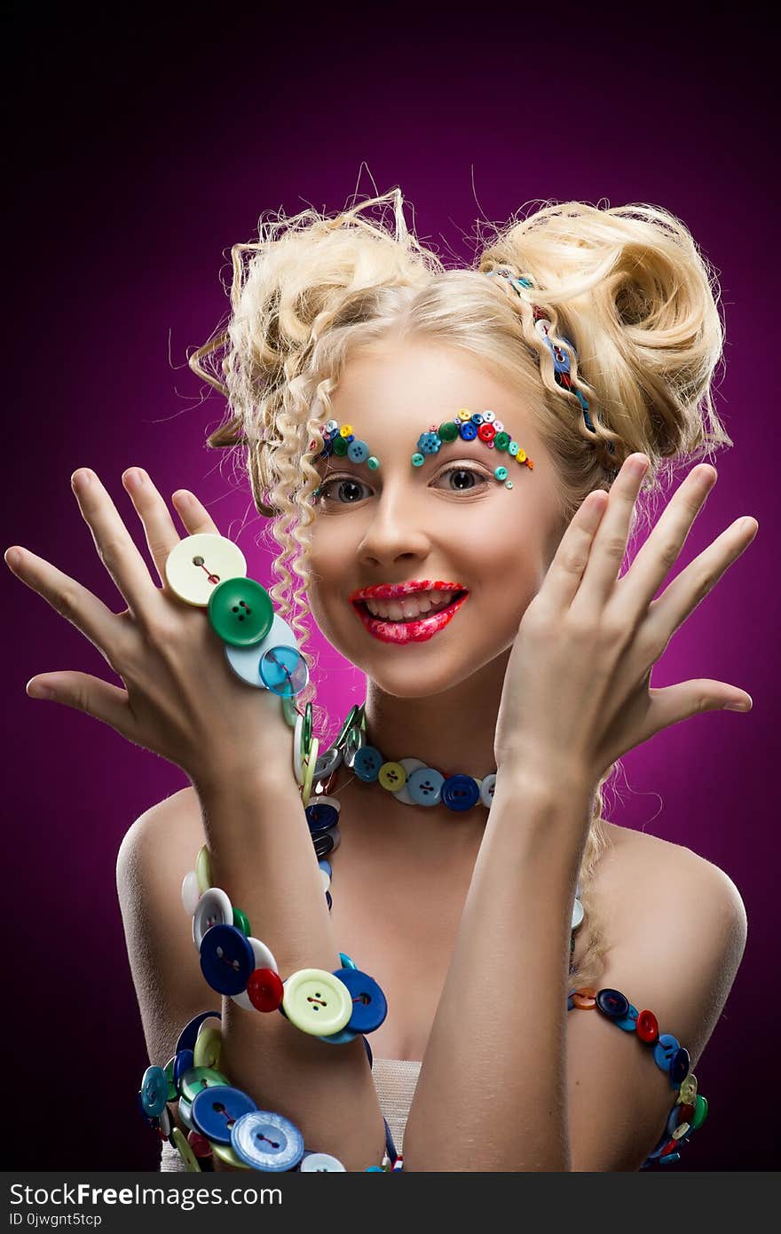 Smiling cute face nice blonde child girl wearing DIY bijou accessories made of multi-colored buttons shows her palms. Funny make-up and pasted buttons on her eyebrows. Pink background. Copy space.
