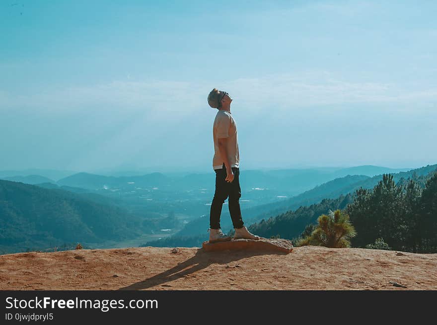 Man Wearing Beige Shirt and Black Pants Looking Up With Blue Sky and Mountains in the Background