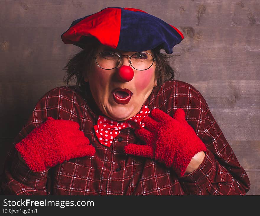 Woman emotional funny cheerful clown with glasses, red nose, shirt, hat and gloves.Wooden background. Woman emotional funny cheerful clown with glasses, red nose, shirt, hat and gloves.Wooden background