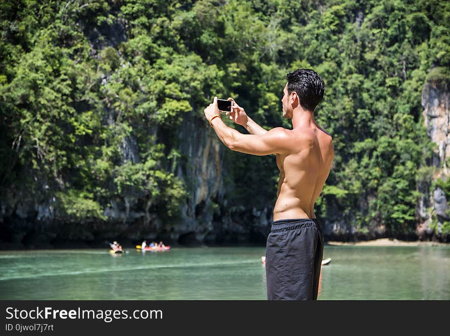 Half body shot of a handsome young man using cell phone, standing on a beach in Phuket Island, Thailand, shirtless wearing boxer shorts, showing muscular fit body. Half body shot of a handsome young man using cell phone, standing on a beach in Phuket Island, Thailand, shirtless wearing boxer shorts, showing muscular fit body