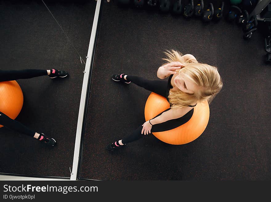 Seductive blonde woman, dressed in dark fitness clothing, posing on large orange exercise ball in front of the mirror. Attractive fitness model posing in position.