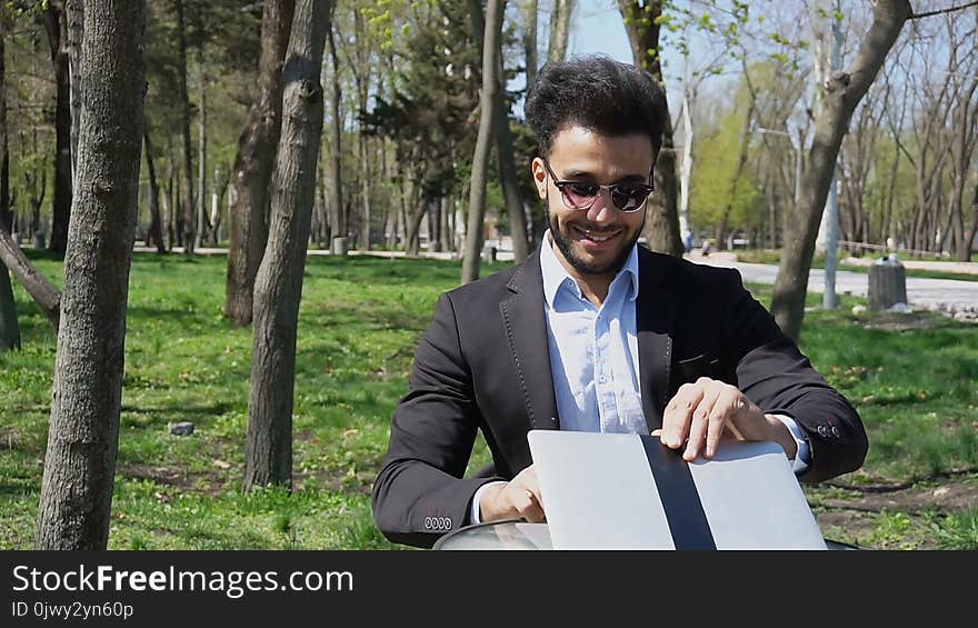 Attractive person sits by table, smiling with dimples on cheeksand uses laptop. Guy has short dark hair, beard, dimples on cheeks and wears sunglasses. Concept of good price of massage modern technologies. Attractive person sits by table, smiling with dimples on cheeksand uses laptop. Guy has short dark hair, beard, dimples on cheeks and wears sunglasses. Concept of good price of massage modern technologies.