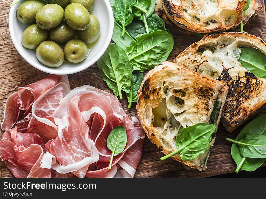 Close up prosciutto, olives, grilled mozzarella spinach sandwiches on wooden background, top view. Mediterranean style snack, appe