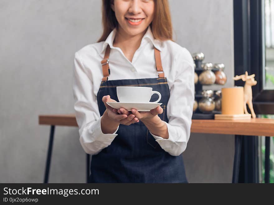 Close up of a young Asian female barista hold a cup of coffee serving to her customer with smile surrounded with bar counter.