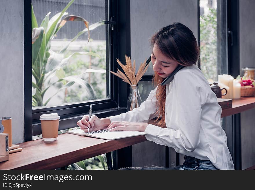 Asian Businesswoman use mobile phone contact customer and writing a note on wood table at coffee shop near window,woking outside