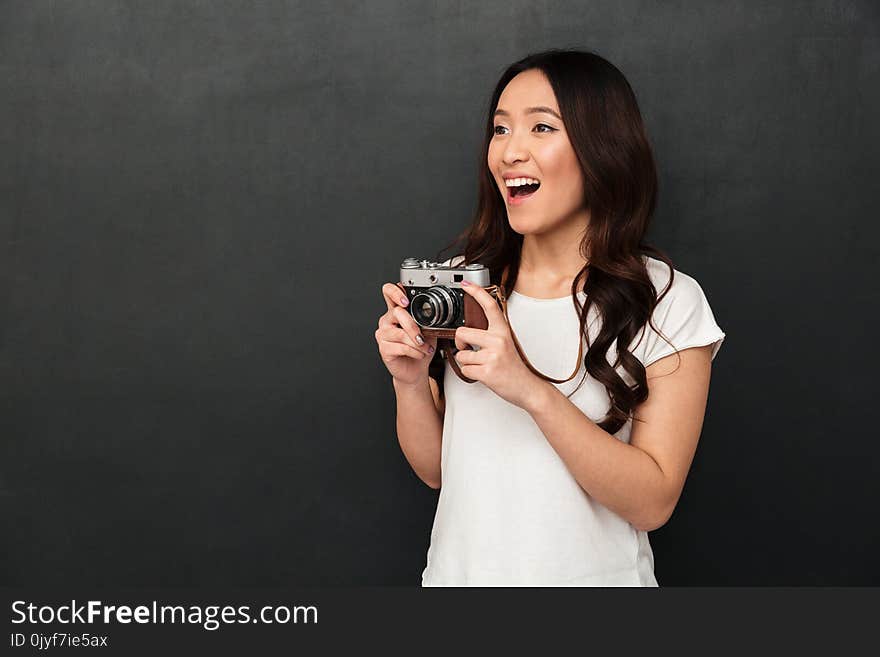 Image of asian young excited woman photographer holding camera isolated over grey wall background. Looking aside.