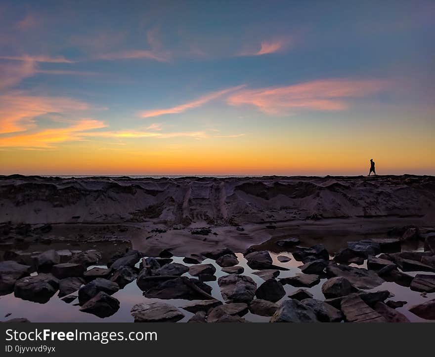 Silhouette Photo of Man Walking on Hill Near Body of Water and Rock Formations