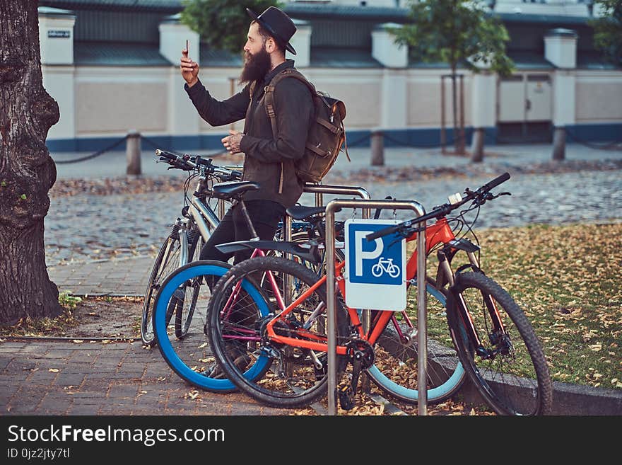 Handsome hipster traveler with a stylish beard and tattoo on his arms dressed in casual clothes and hat, using the phone, standing on the bicycle parking. Handsome hipster traveler with a stylish beard and tattoo on his arms dressed in casual clothes and hat, using the phone, standing on the bicycle parking.