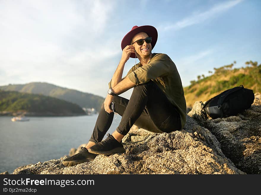 Nice smiling guy in sunglasses is listening music while sitting on the rocky cliff on the sunny background of the sea bay. He wears dark jeans and sneakers, olive shirt and crimson hat. Horizontal. Nice smiling guy in sunglasses is listening music while sitting on the rocky cliff on the sunny background of the sea bay. He wears dark jeans and sneakers, olive shirt and crimson hat. Horizontal.