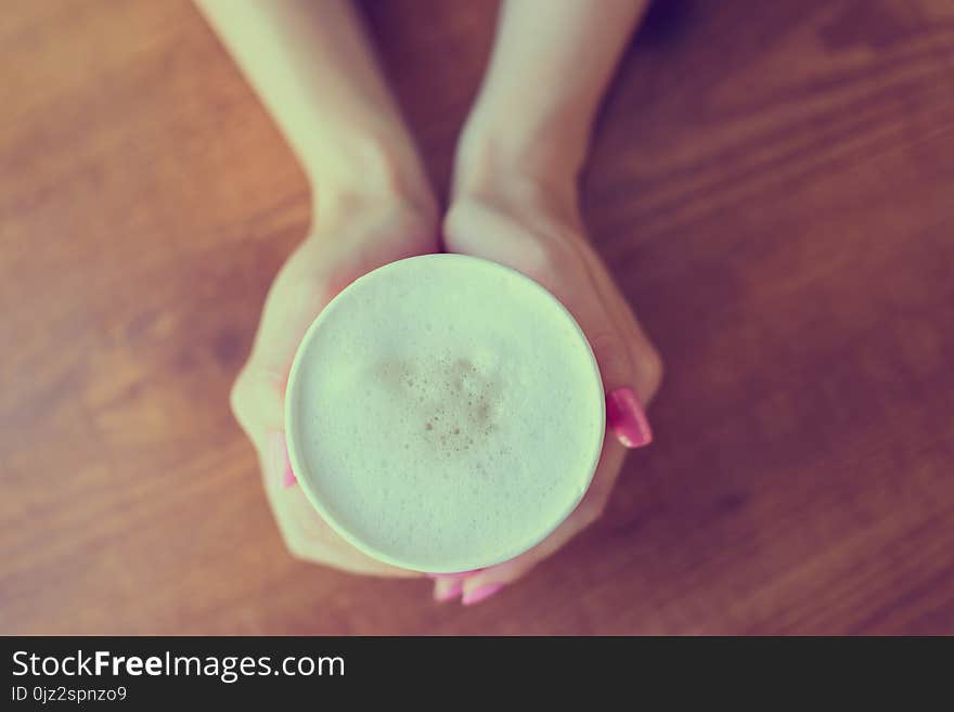 Woman`s hands holding a cup of frothy coffee over wooden background, top view