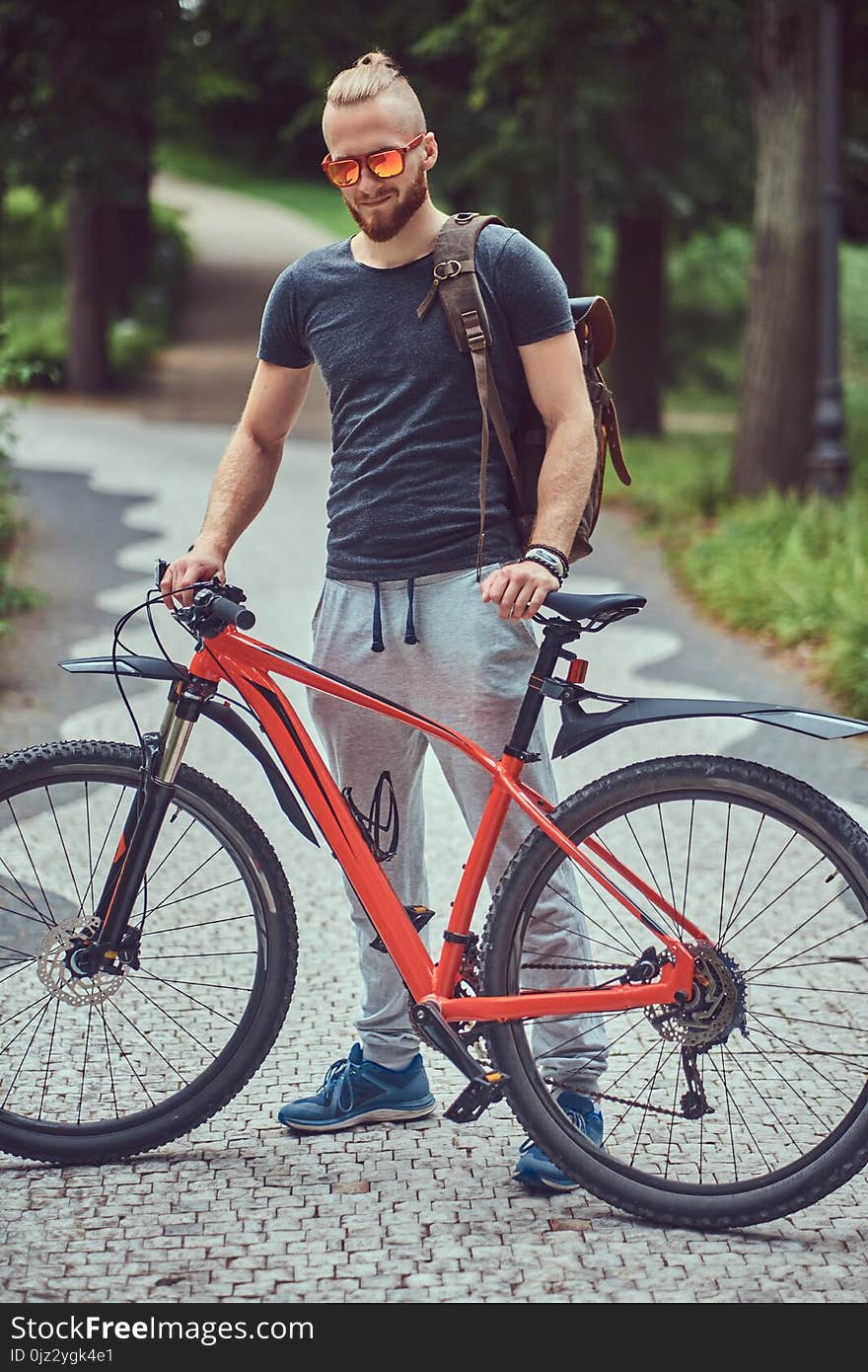 Handsome redhead male with a stylish haircut and beard dressed in sportswear and sunglasses walks in the park with a bicycle and backpack. Handsome redhead male with a stylish haircut and beard dressed in sportswear and sunglasses walks in the park with a bicycle and backpack.