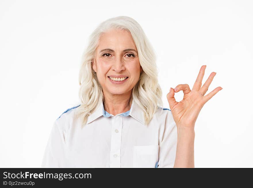 Image of mature old smiling woman standing isolated over white background looking camera showing okay gesture. Image of mature old smiling woman standing isolated over white background looking camera showing okay gesture.