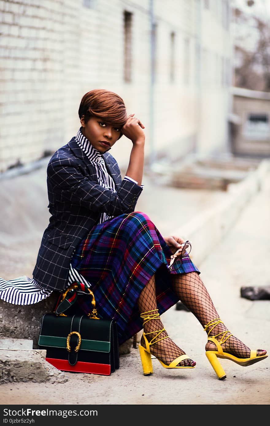 Woman Wearing Black and Grey Tattersall Blazer and Multicolored Plaid Skirt With Black Mesh Stocking and Yellow Chunky Heeled Sandals Sitting on Grey Concrete Pathway