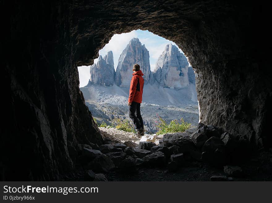 Man in Red Jacket Standing Outside of Cave in Front of Three Mountains