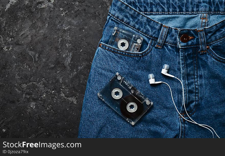 Jeans, audio cassette, headphones layout on a black concrete table. Conceptual photo illustrating the fascination of listening to music. Top view.