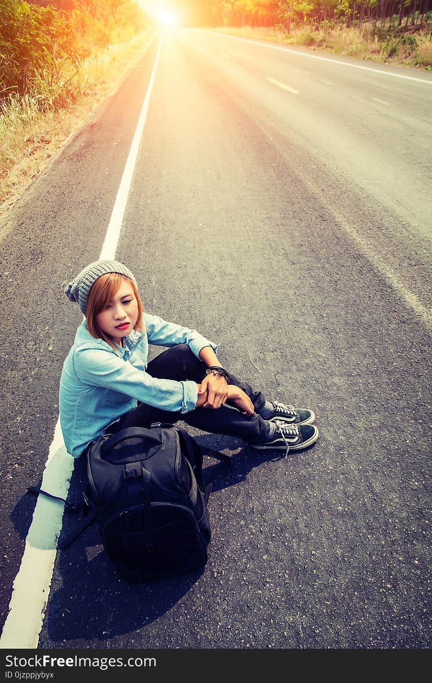 Rear view of a young woman hitchhiking carrying backpack sitting on the road