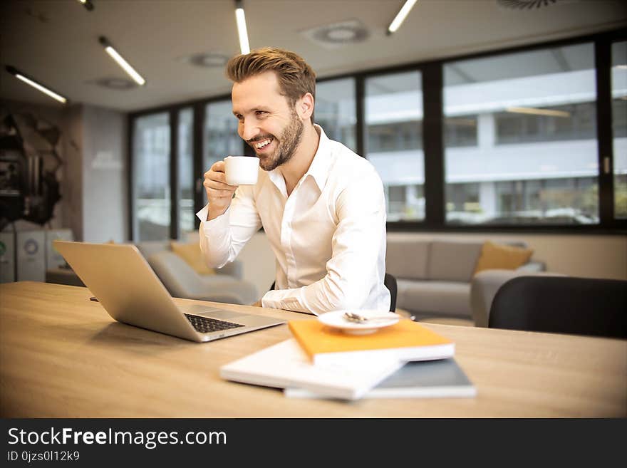 Depth of Field Photo of Man Sitting on Chair While Holding Cup in Front of Table