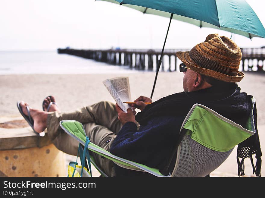 Man Sitting on Chair Under Blue Umbrella Near Beach