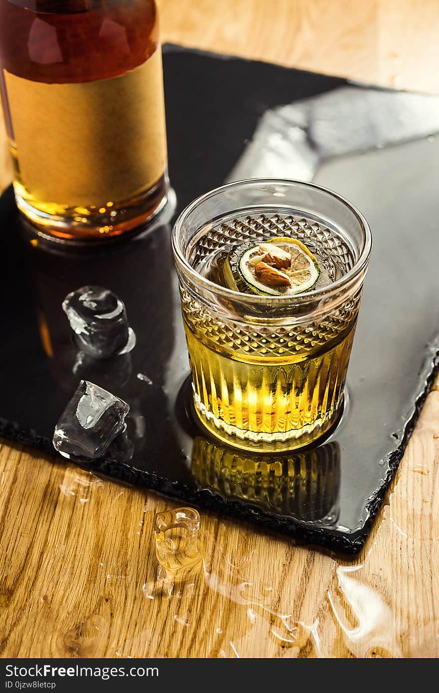 Glass of whiskey cognac or brandy with lemon and ice cubes standing on the bar counter with a bottle on the background