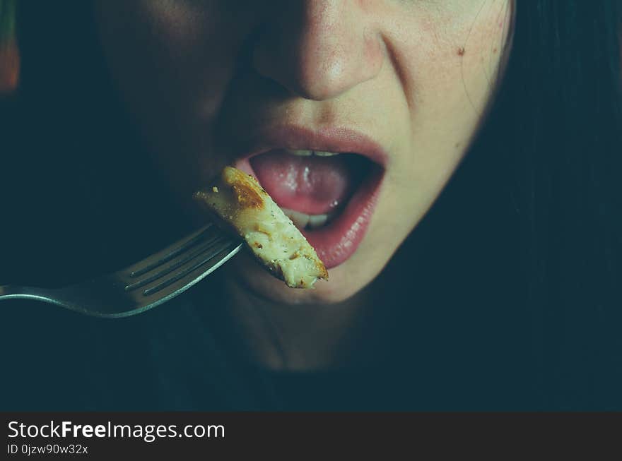 Part of the girls face with mouth and tongue holding fork with chicken meat eating in the night, hiding from other people, illuminated with light from the fridge, obesity concept, selective focus.