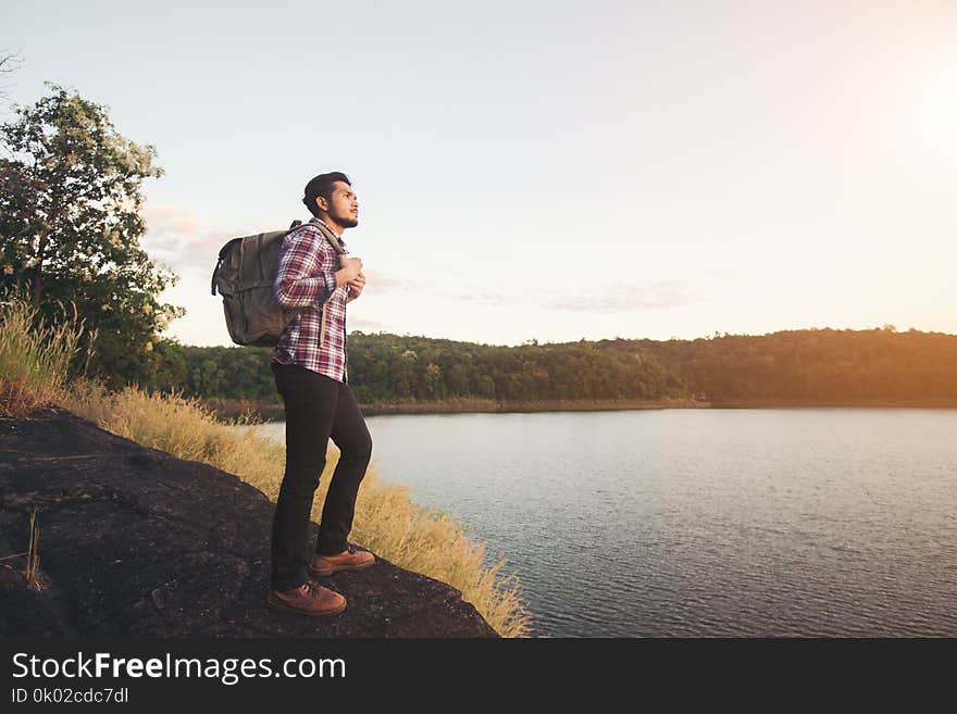 Hipster hiker man standing on the rock and enjoying sunset over lake.