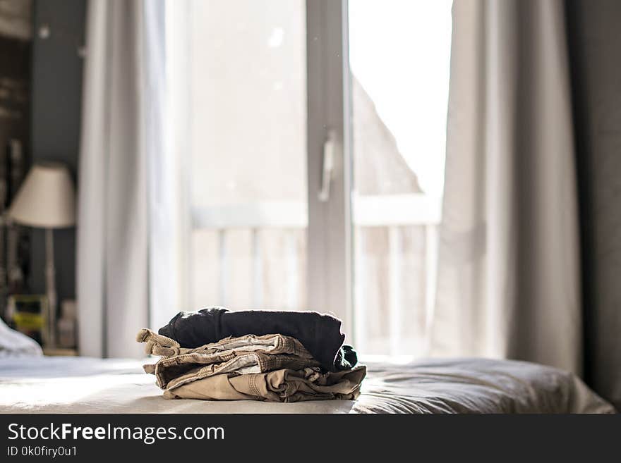 Pile of clothes on bed beside window. Curtains behind and natural light. Pile of clothes on bed beside window. Curtains behind and natural light.