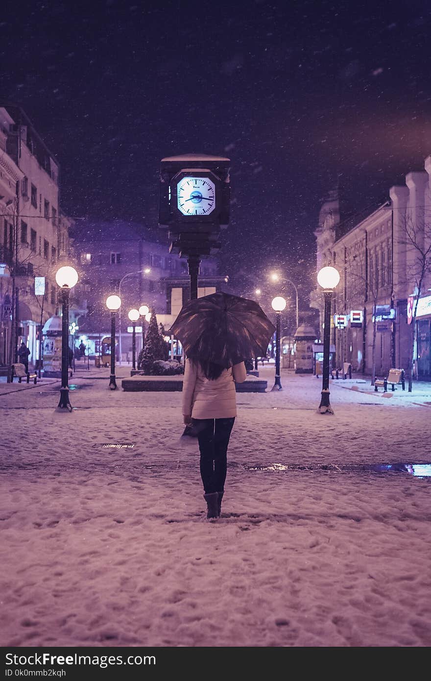 Person in White Long-sleeved Top and Black Pants Under Black Umbrella Standing Near Street Clock