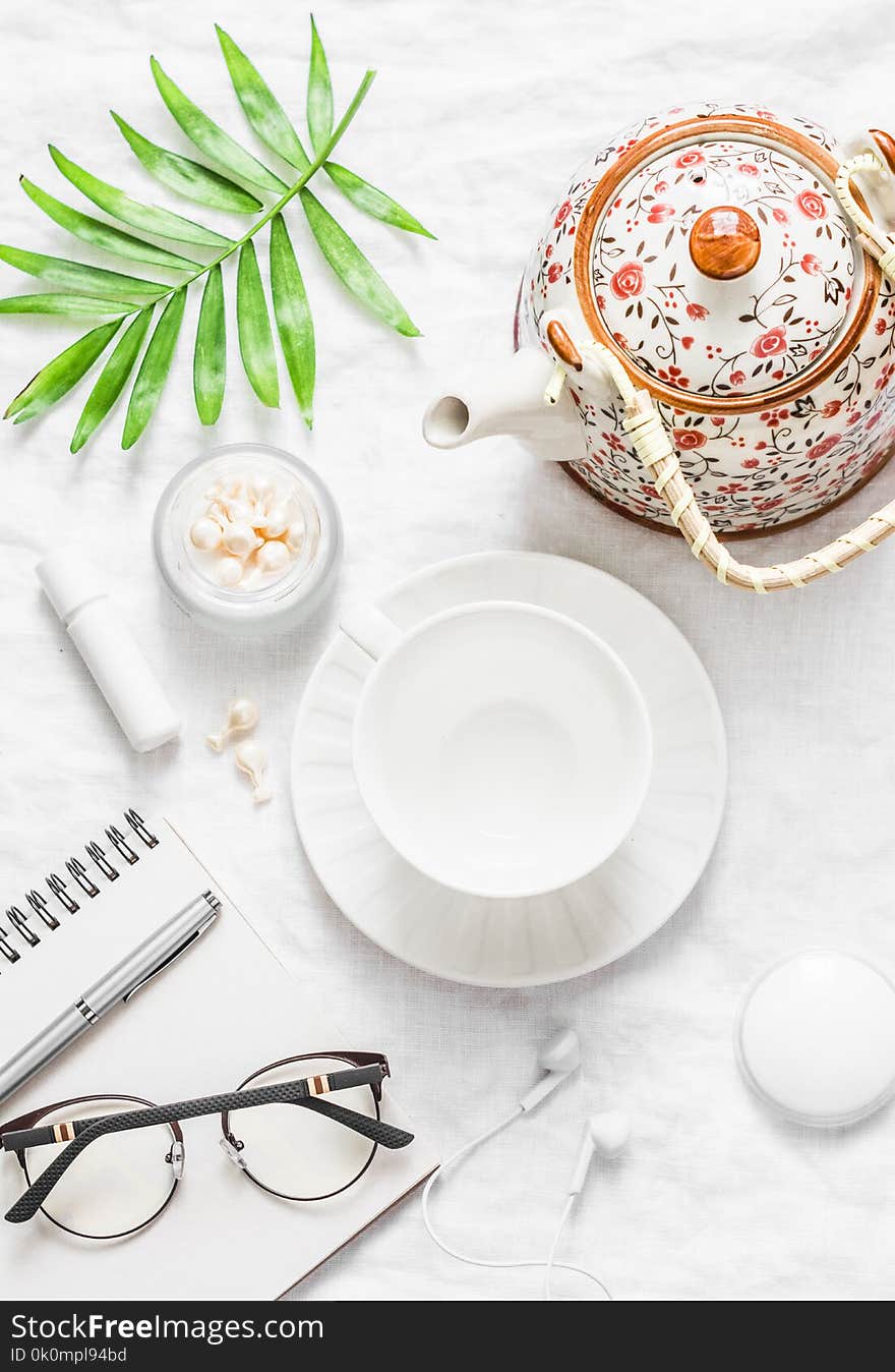 Flat lay cozy morning inspiration planning female table - teapot, tea cup, notepad, glasses, lipstick, face cream, green leaf flower on light background, top view
